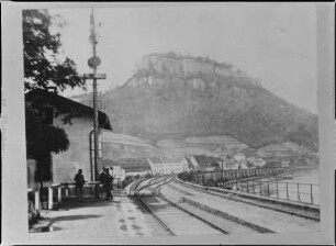 Königstein, Bahnhof. Böhmischer Bahnsteig. Ansicht mit drei Personen um einen Fahnen-/Signalmast gegen den Königstein mit Festung