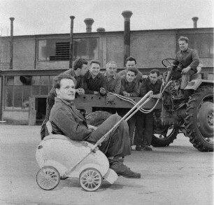 Schönebeck. VEB Traktorenwerk Schönebeck. Produktionsbereich Geräteträger RS 09. Gruppe Kraftfahrzeugschlosser an und auf einem Fahrzeug auf dem Werkgelände, davor ein Schlosser in einem Kinderwagen sitzend