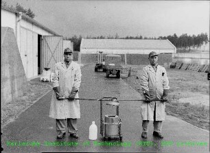 Zwei Mitarbeiter beim Transport von Abfallbehältern für radioaktives Material auf dem Gelände des Kernforschungszentrums Karlsruhe (KfK)