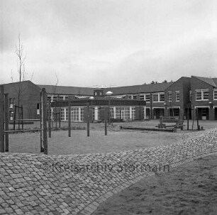 Bargteheide: Kreisgymnasium II [später Gymnasium Eckhorst]: Eckhorst: Außenaufnahme mit Uhr: davor Schulhof, Grünflächen, Sandfläche mit Turngeräten, vorne Erdwall mit Pflasterung