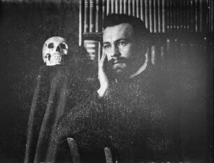 Portrait Rudolf Zimmermann : Portrait von Rudolf Zimmermann neben einem menschlichen Schädel vor einem Bücherregal.