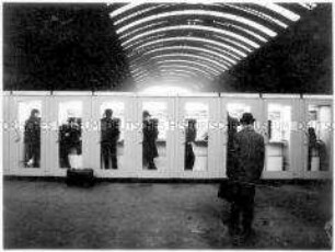 Reisende in aufgereihten Telefonzellen einer Bahnhofshalle