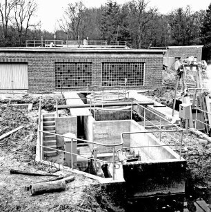Abwasser: Klärwerk: Wöhrendamm: 3. Bauabschnitt: Schmutzwassereinläufe: Becken im Rohbau mit Geländer und ersten Einbauten: rechts Förderband: hinten Maschinen- und Betriebsgebäude im Rohbau: im Hintergrund Bäume