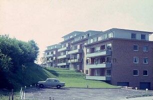 vorn Parkplatz mit Pkw: dahinter Grünflächen mit Büschen und Wohnblocks
