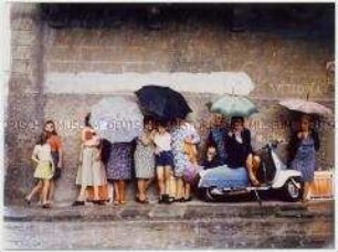 Frauen in bunten Kleidern und Kinder stehen mit aufgespannten Regenschirmen an einer Mauer, es regnet