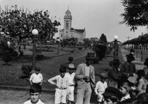 Gruppe am Platz in Bica de Pedra : Eine Gruppe Männer und Kinder stehen an einem als Grünanlage gestalteten Platz mit Bäumen und Sträuchern in Bica de Pedra. Im Hintergrund die Kirche des Ortes.