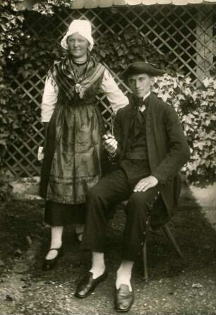 Ehepaar in klassischer Pose. Mann mit Pfeife sitzend auf Hocker. Frau daneben stehend, in Volkstracht gekleidet.Halbtotale