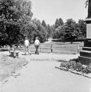 Alter Friedhof: vorn rechts Ehrenmal: links Bank: daneben zwei Spaziergänger: hinten Mitte Grünanlage mit Baumbestand