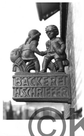 Nasenschild Bäckerei H. Schriefer: Bad Oldesloe: Knabe und Mädchen bei Händedruck, mit Korb, Brot, Getreidegarben: an Hauswand;