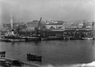 Hafen : Hafendock mit Schiffen, Booten und Kränen, dahinter Häuser einer Stadt.