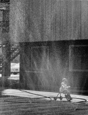 Sommer. Ein kleines Mädchen auf dem Dreirad erhält eine Dusche vom Rasensprenger