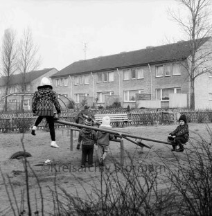 Kinderspielplatz für Siedlung Gartenstadt: Kieler Straße: Kinder auf den Wippen: dahinter Klettergerüst: Jägerzäune, Sitzbank, Baum: Fußweg: hinten Gartenfront der Reihenhäuser an Jahnstraße