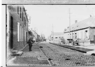 Straßenszene in einer Ortschaft, mit Pferdefuhrwerken und Soldat