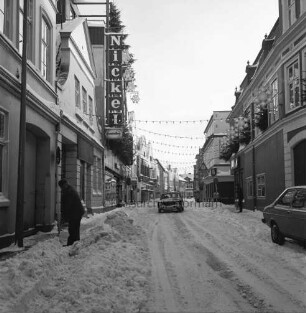Mühlenstraße: vorn links und hinten rechts Männer beim Schneeschippen: links Kaufhaus Nickel: rechts Feinkosthandlung Petersen mit Weihnachtsschmuck: davor parkender Pkw: dahinter Abzweigung der Hindenburgstraße: hinten Mitte Pkw: Winter