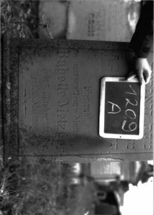 Grabstein 1209 (Index: 100104381) Material: Sandstein Zustand: einige Abbrüche und Risse, Inschrift noch gut erhalten Maße HxBxT: 145x50x16 cm Beschreibung: aufgesockelte Stele mit Giebelaufsatz, der mit eingeritzten Ranken geschmückt ist, umlaufende Auskragungen oberhalb des Schriftfeldes, das durch eine eingeritzte, umlaufende Rille abgeteilt ist: Grabstein 1209A