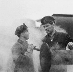 Zwei Eisenbahner zwischen Gleisen im Gespräch, umgeben von Dampfschwaden (einer Lokomotive), im Vordergrund ein Hülsenpuffer (Serie Deutsche Reichsbahn - Brigade "Otto Buchwitz")