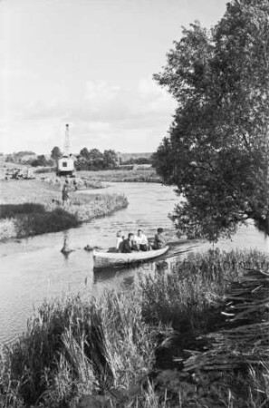 Trave: Flussregulierung: vorn Uferböschung mit Schilf: rechts Baum: in der Mitte Motorboot mit vier Personen im Wasser: hinten links Mann, Bagger, Loren und Baumaterial: hinten Wiesen und Büsche: dahinter Häuser von Sehmsdorf zwischen Bäumen