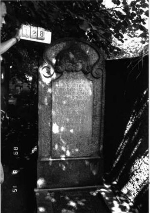 Grabstein 128 (Index: 100105327) Material: Granit Zustand: verwittert und gebrochen Maße HxBxT: 177x67x23 cm Beschreibung: Stele, Rundbogenabschluß nach unten in Voluten auslaufend, erhabenes Schriftfeld Symbolik: Voluten, Blattwerk über Schriftfeld: Grabstein 128