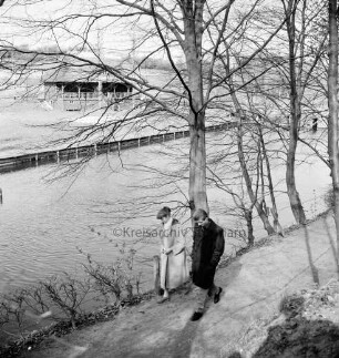 Trave: vorn Wanderweg mit Baumreihe am Ufer: vorn Paar: hinten links am gegenüberliegenden Ufer Badeanstalt: hinten rechts Brücke