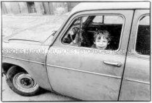 Kleiner Junge guckt aus einem am Straßenrand stehenden alten Auto (Sonderthema: Straßen-Kinder)