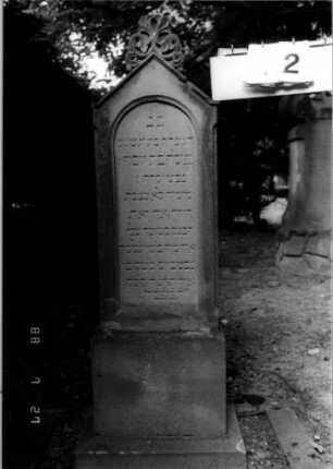 Grabstein 12 (Index: 100110217) Material: Sandstein Zustand: Stein leicht brüchig Maße HxBxT: 128x38x20 cm Beschreibung: Stele, getreppter Sockel, giebelartiger Abschluß mit kleiner Verdachung und Mittelakroter, eingetieftes Schriftfeld mit Rundbogen Symbolik: ornamentales Mittelakroter: Grabstein 12