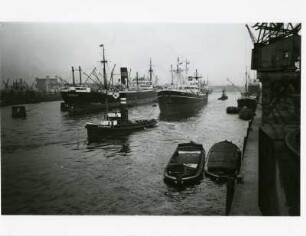 Hamburg. Betriebsamkeit im Hafen. Ein Frachtschiff wird vom Schlepper "Steinwerder" an seinen Liegeplatz verbracht. Andere Schiffe liegen auf Reede bzw. an den Kaianlagen