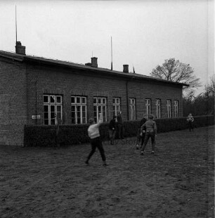 Volksschule: davor Hecke: vorn Schulkinder beim Spiel auf Schulhof