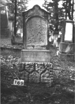 Grabstein 169 (Index: 99899021) Material: Sandstein Zustand: stark verwittert Maße HxBxT: 140x54x25 cm Beschreibung: aufgesockelte Stele mit Konvexbogenabschluss, eingeritzte Mittelpalmette und Voluten, unterer Sockel felsartig gearbeitet Symbolik: Palmette, Voluten: Grabstein 169