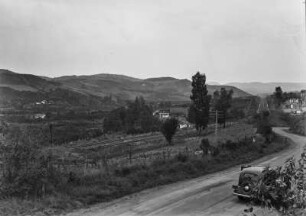 Landschaftsblick : Blick über Plantagen auf den Hängen und einzelnen Gebäuden an auf der linken Bildseite. In der Mitte verlaufen zwei Gleise, rechts eine Siedlung mit Gebäuden.