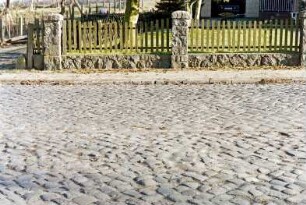 Dorfstraße: Kopfsteinpflaster, Straßenmitte: hinten Fußgängerweg, Gartenzaun, links Einfahrt