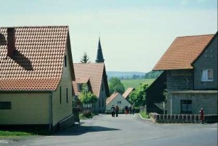 Dorfstraße und Wohnhäuser mit neu gedeckten Dächern; hinten Kirchturmspitze