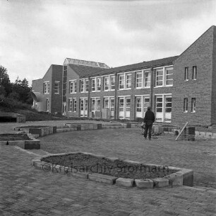 Bargteheide: Kreisgymnasium II [später Gymnasium Eckhorst]: Eckhorst: vor Fertigstellung: Herrichtung der Außenanlagen: Arbeiter beim Pflastern des Halbrunds mit Treppen und Anpflanzungen, Schubkarre: links Lüftungsrohr