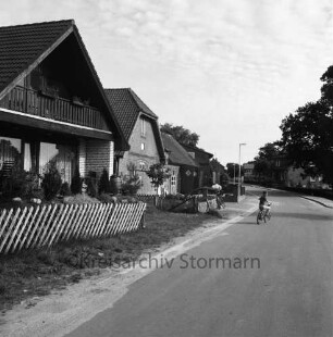 Dorfstraße: links und hinten rechts Einfamilien- und Bauernhäuser mit Vorgärten: rechts Kind mit Fahrrad