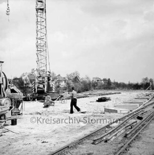Bahnhofsgelände: Lagerschuppen der Claudius-Mühle in Bau: Fundamente, ein Arbeiter: Baukran auf Schienen, an Haken Betonkübel, links Betonmaschine: im Vordergrund Gleis, Schienenstücke: hinten Bauschild: im Hintergrund Häuser an Bischofsteicher Weg