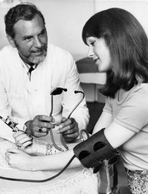 Beim Vertrauensarzt. Einer Patientin wird der Blutdruck gemessen. Sie liest die Werte selbst ab.