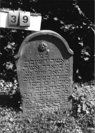 Grabstein 39 (Index: 99796345) Material: Sandstein Zustand: unten abgeblättert mit Textverlust Maße HxBxT: 78x47x11 cm Beschreibung: Stele, schräg eingezogener Rundbogenabschluß mit Verdachung Symbolik: Blüte: Grabstein 39