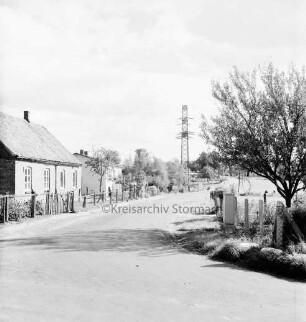 Dorfstraße: rechts Zaun, Baum, Verteilerkasten: dahinter Weide: links Häuser mit Vorgärten und Zaun: hinten Mitte Strommast