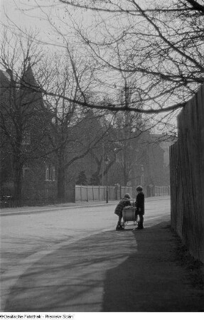 Zwei Kinder mit Kinderwagen auf dem Bürgersteig einer leeren Straße