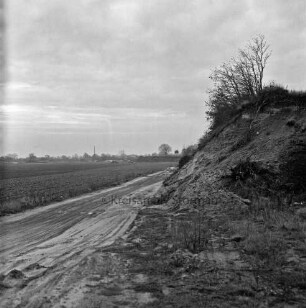 Feldmark: Wirtschaftsweg: rechts Hang von Kiesgrube mit Buschwerk: hinten links Sandhaufen, Lkw und Schornstein hinter Bäumen