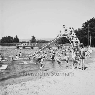 Freibad Poggensee: Badegäste am Sandstrand und im Wasser, vorne eine Rutsche, im Hintergrund der Badesteg
