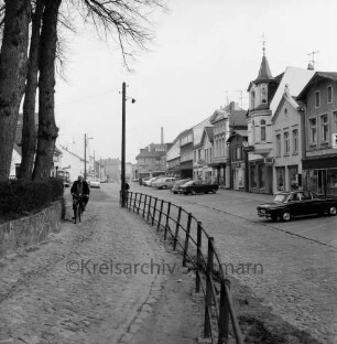 Paul-von-Schoenaich-Straße: Blick Richtung Nordosten: links Kirchsteig, Kirchhofmauer, Bäume, Fahrradfahrer: Bildmitte Trenngeländer, Strommast mit Straßenlampe: rechts vor Ladenzeile Parkplätze, parkende Autos