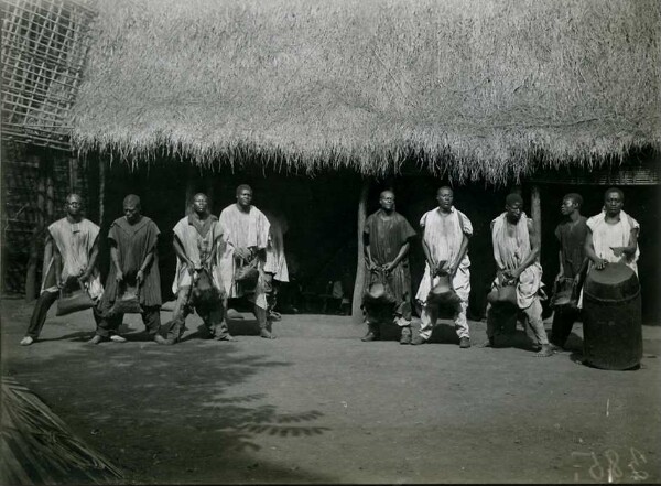 Danse de huit hommes, debout quatre par quatre les uns à côté des autres, tenant un sac à la main. Un neuvième homme joue du tambour.