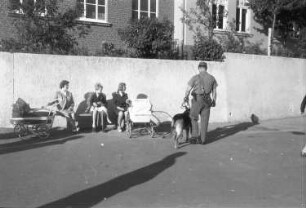Berlin (?). Blick von der westdeutschen Seite auf die Mauer. Davor sitzen Passanten mit Kinderwagen, ein Grenzschützer mit Hund läuft Streife