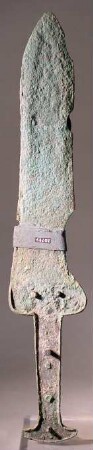 Kurzes breites Dolchmesser. Klinge in zwei Teile gebrochen, auch sonst beschädigt. Auf der einen Seite am Übergang von Griff und Klinge und weiterhin auf dieser selbst Reste von Holz, von der Verschalung des Griffes und von der Scheide (?)