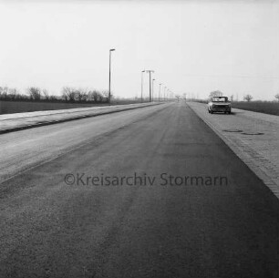 Gewerbegebiet West: asphaltierte Straße mit Parkstreifen, Fußwegen und Straßenlampen, rechts Pkw: dahinter Bäume