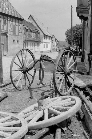 Ansicht einer Dorfstraße, im Vordergrund Wagenräder