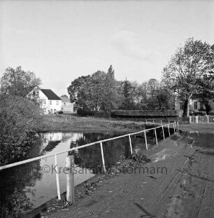 Alte Dorfstraße: links Teich: davor Zaun: hinten Häuser mit Gärten und Hecken zwischen Bäumen