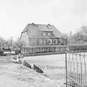 Volksschule: Schulgebäude hinter Hecke: im Vordergrund vertiefte Rasenfläche: rechts Metallzaun