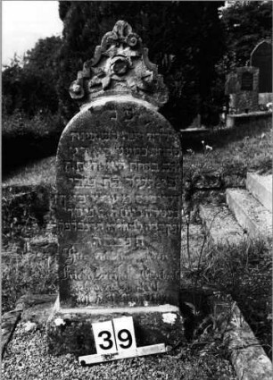 Grabstein 39 (Index: 99801986) Material: Sandstein Zustand: verwittert mit Textverlust Maße HxBxT: 92x33x14 cm Beschreibung: Stele, Sockel, Rundbogenabschluß mit großem Mittelakroter Symbolik: Blüte und Blätter im Mittelakroter Besonderheit: Kindergrab: Grabstein 39