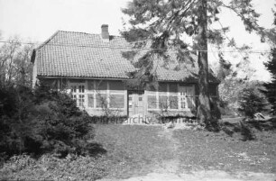 Kneeden: Forsthaus: davor Sandhaufen: links auf Bank Person: davor Büsche: rechts großer Baum: hinten weitere Bäume